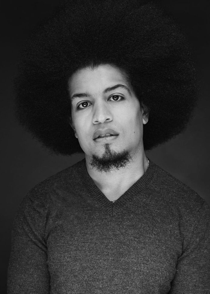 Black and white portrait of a man with a large afro and goatee, wearing a dark v-neck sweater.