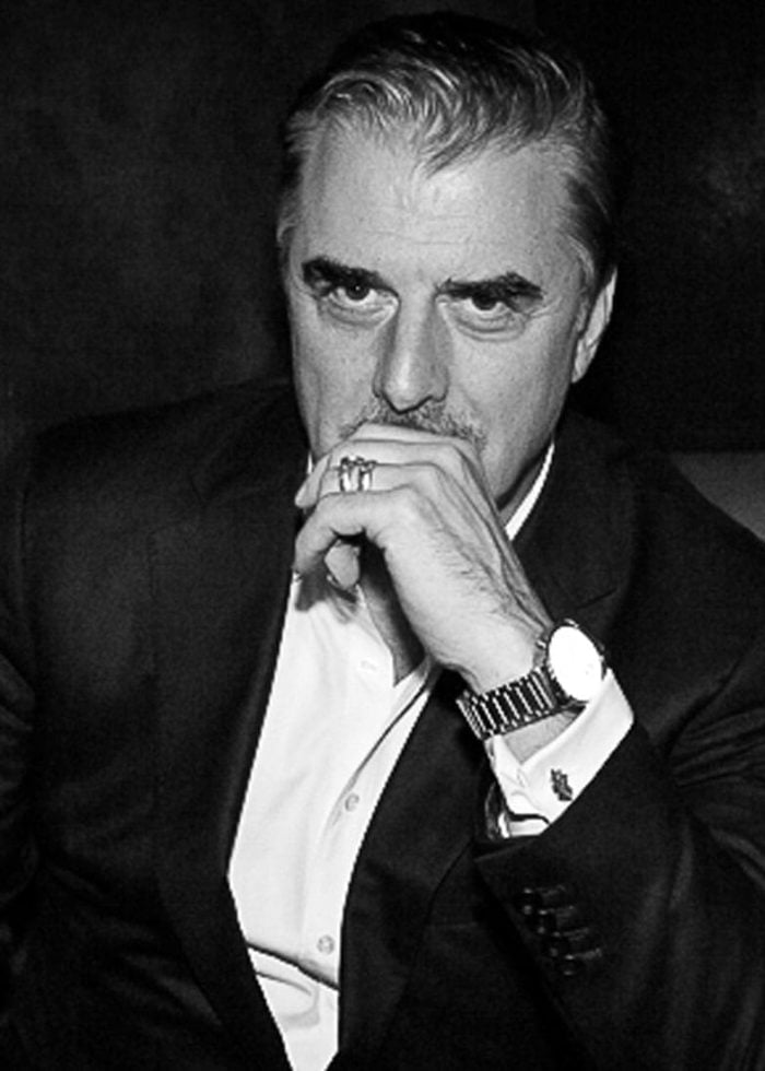 Chris Noth in a black suit, looking thoughtful, wearing a watch and ring.