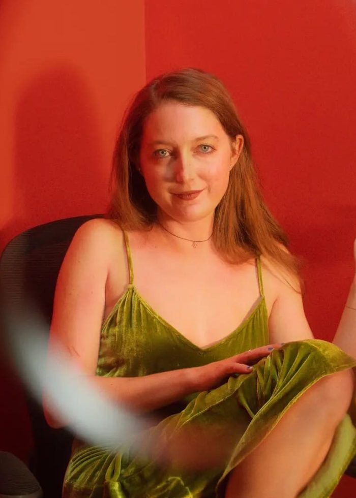 Clare Barron in a green velvet dress, seated in a red-walled room.