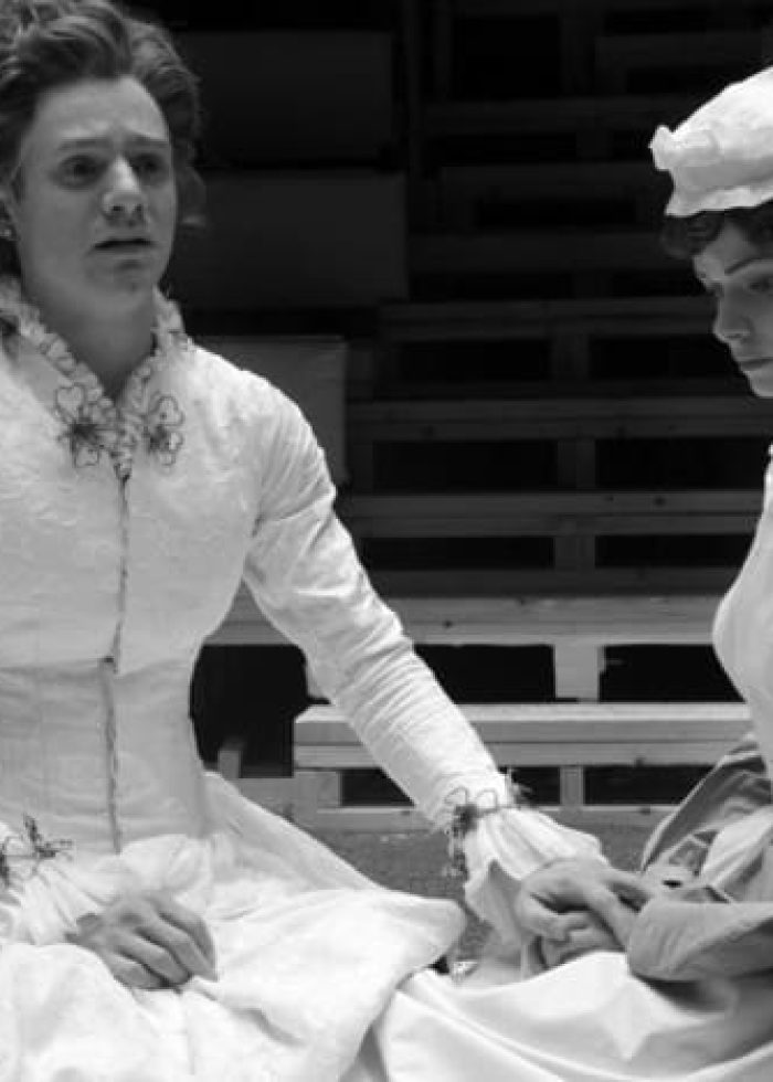 Actors in a scene from Cloud Nine, one in a white dress, the other in a maid's uniform, holding hands.