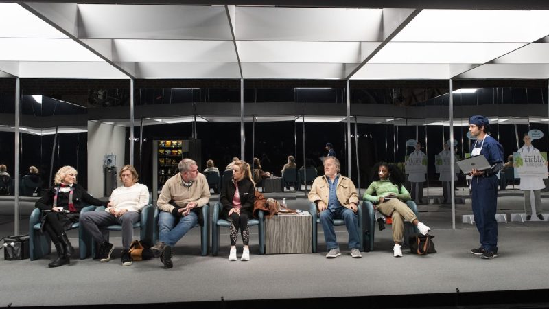 People waiting in a modern medical office, possibly related to "I'm Revolting." A doctor holds a clipboard.