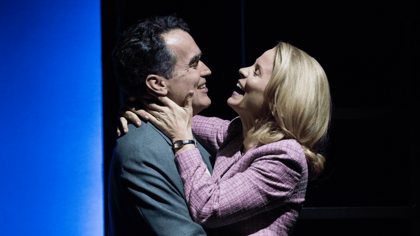 A man and woman embrace, laughing, in a dimly lit setting.