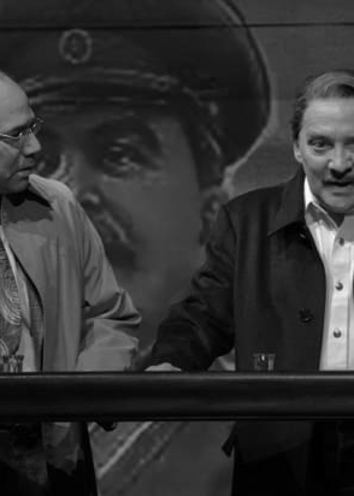 Two actors in a theater production, possibly "Describe the Night," standing behind a bar with a historical backdrop.