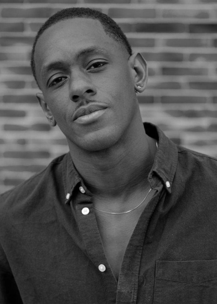 Stylish man with short hair posing in front of a brick wall, wearing a dark shirt.