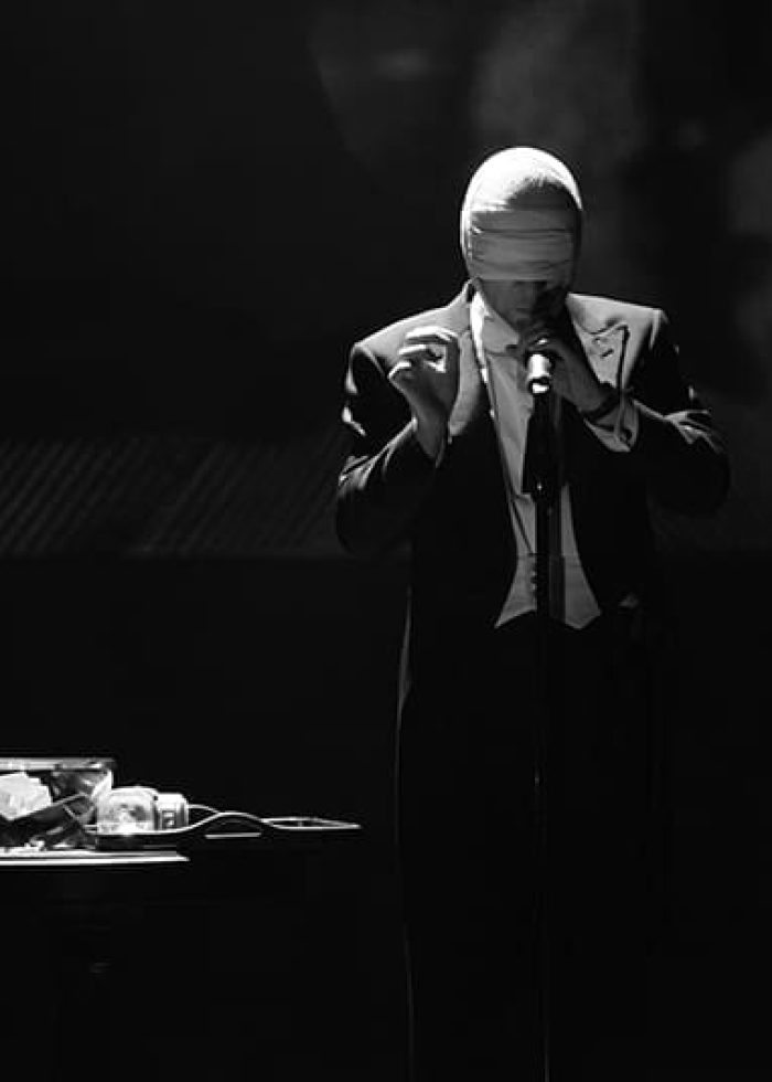 Derren Brown performs on stage, blindfolded, in a suit. A table with objects is in the foreground.