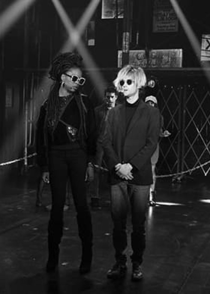 Two people in sunglasses stand on stage in a gritty, industrial setting. Performers are in the background.