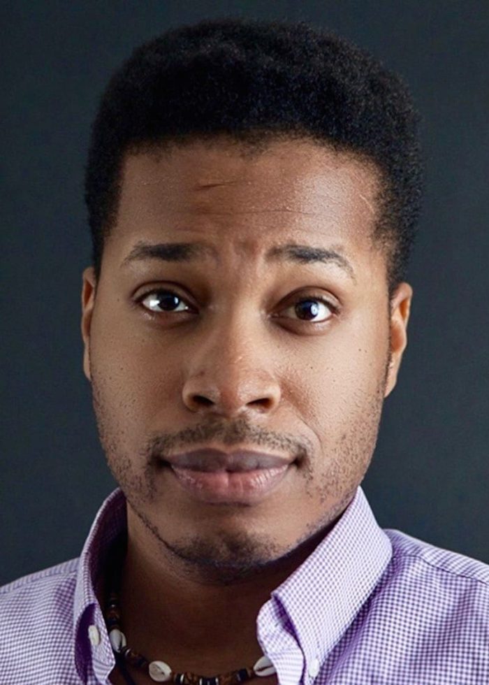 Close-up portrait of Dwayne Gayle wearing a purple checkered shirt and necklace, looking directly at the camera.