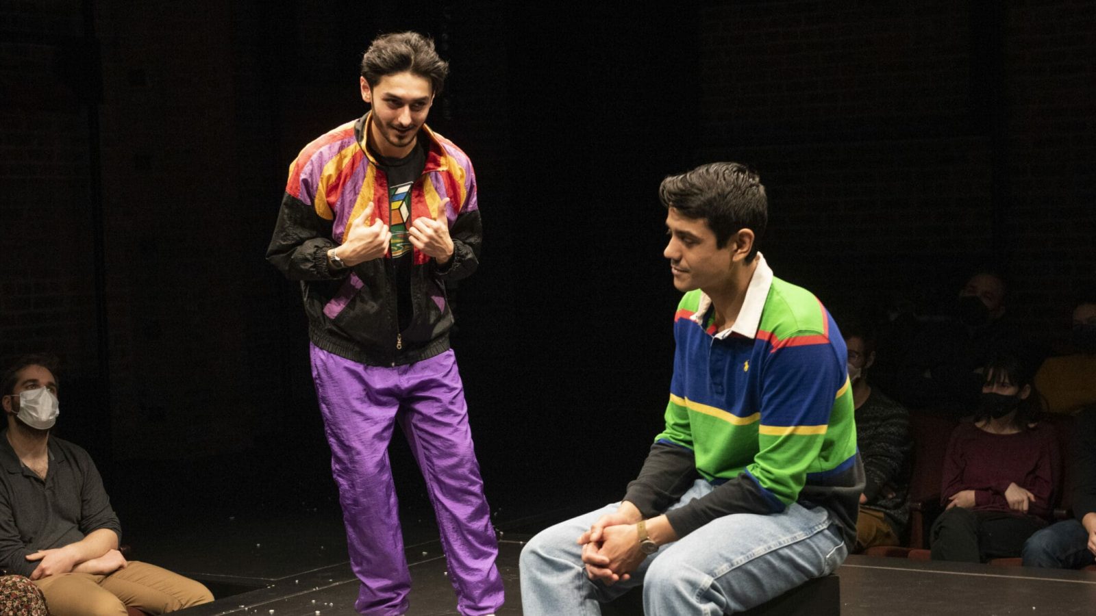 Two actors in a theatrical performance, one in a vibrant jacket and purple pants, the other in a striped shirt and jeans. Audience members wear masks.