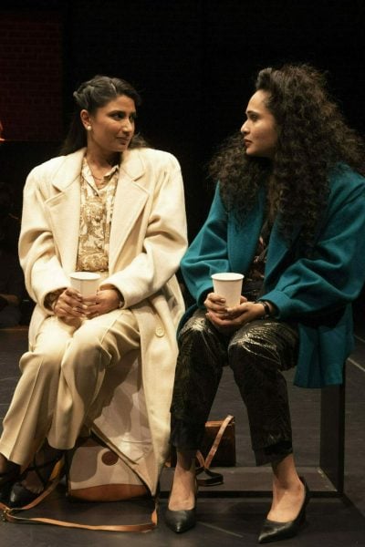 Two women in coats sit on stage, holding coffee cups, during a performance of Elyria.