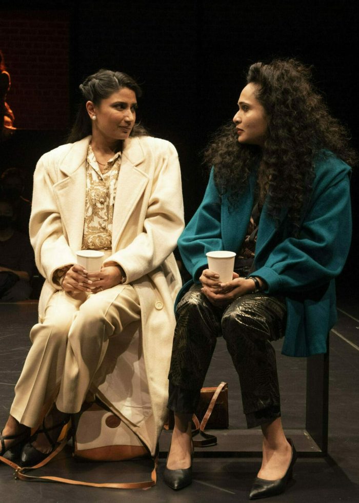 Two women in coats sit on stage, holding coffee cups, during a performance of Elyria.