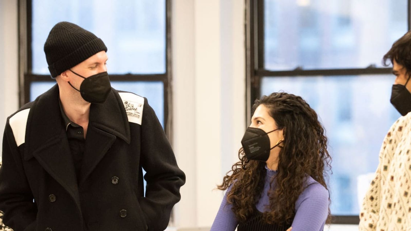 Four masked individuals, two men and two women, converse in a bright room.