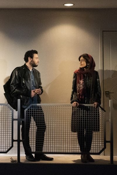 Man and woman converse on a balcony, separated by a metal railing.