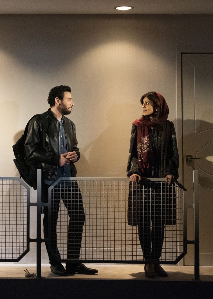 Man and woman converse on a balcony, separated by a metal railing.