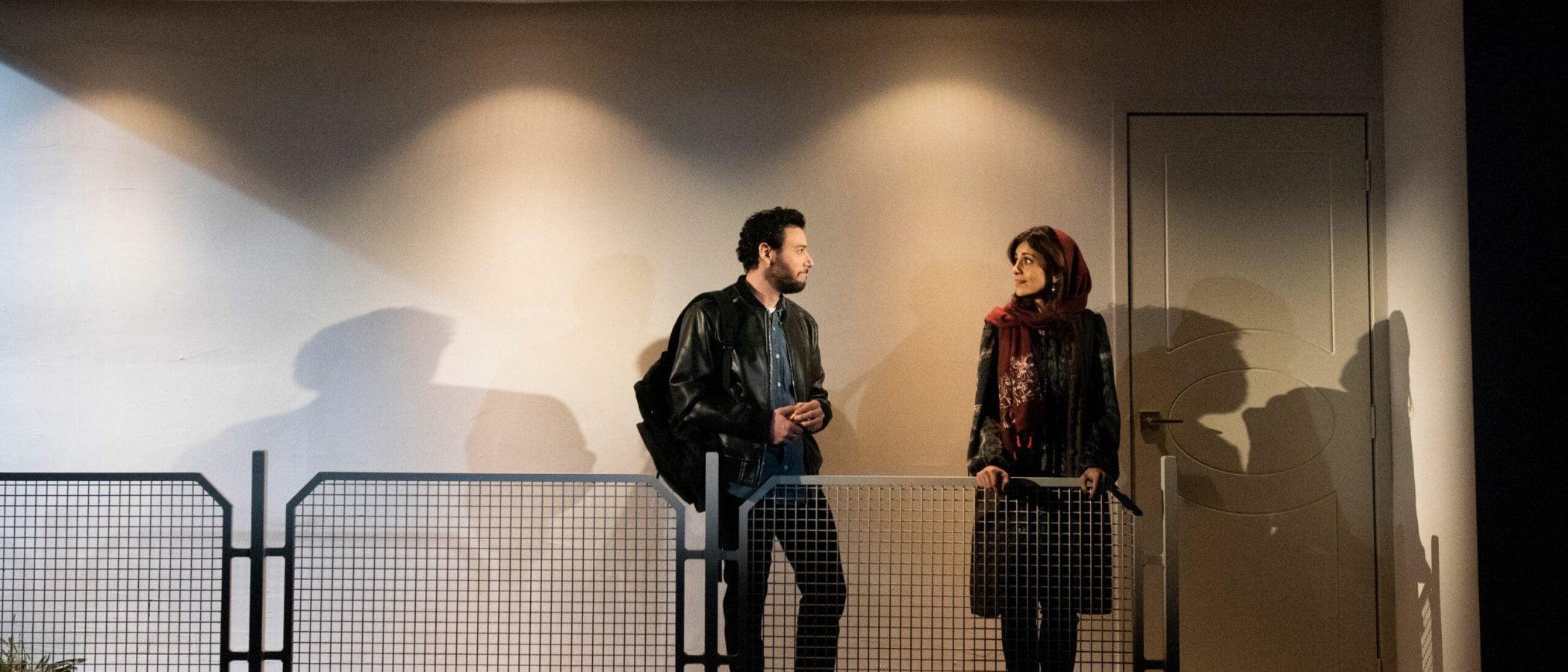 Man and woman converse on a balcony, separated by a mesh fence.