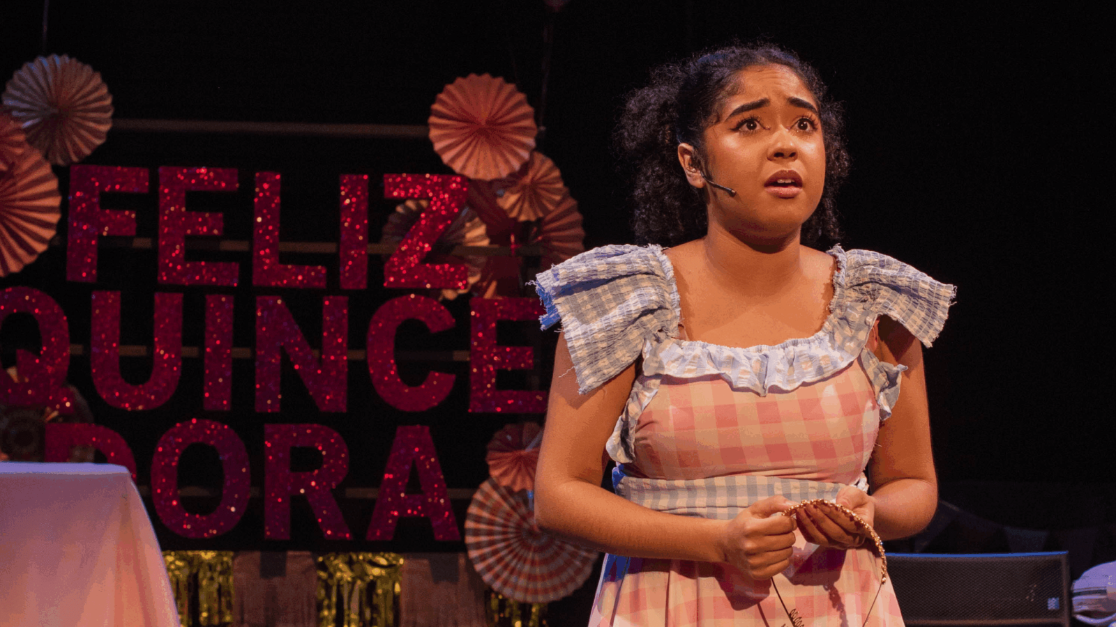 Young actress in a pink gingham dress performs during El Otro Oz play, with "Feliz Quinceañera" sign in the background.
