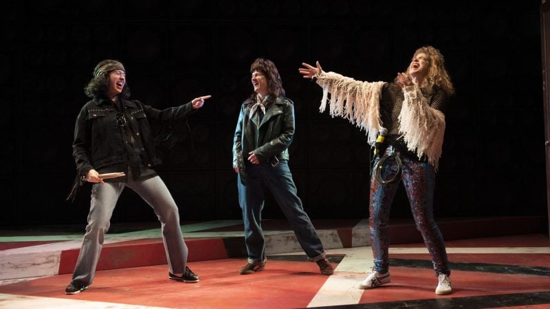 Three actors on stage in 70s rock costumes, performing a scene.