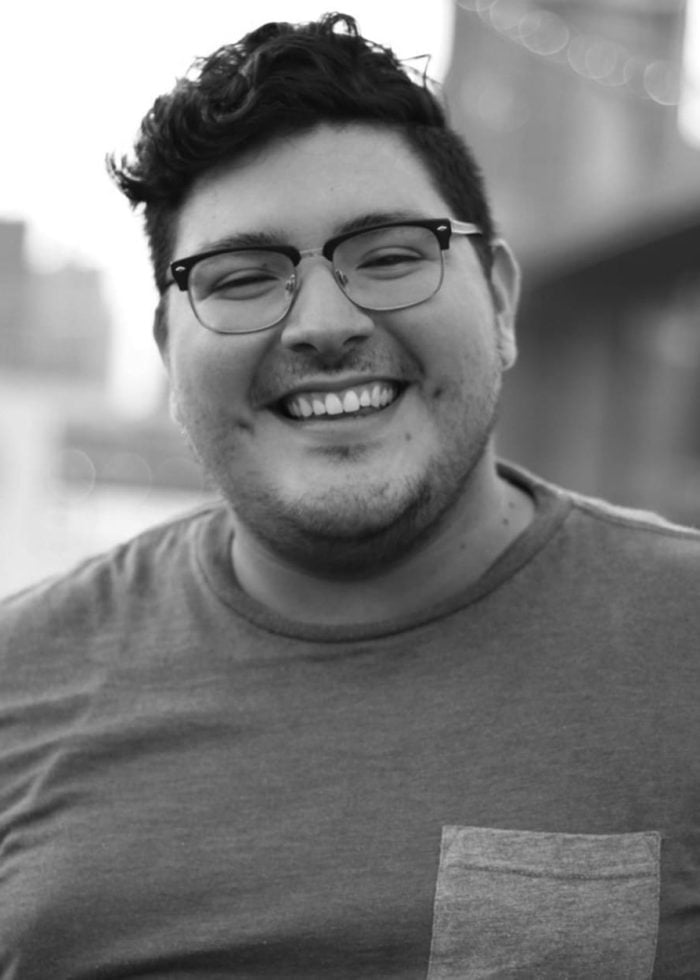 Eli Gonzalez smiles, wearing glasses and a grey shirt, in a black and white photo.