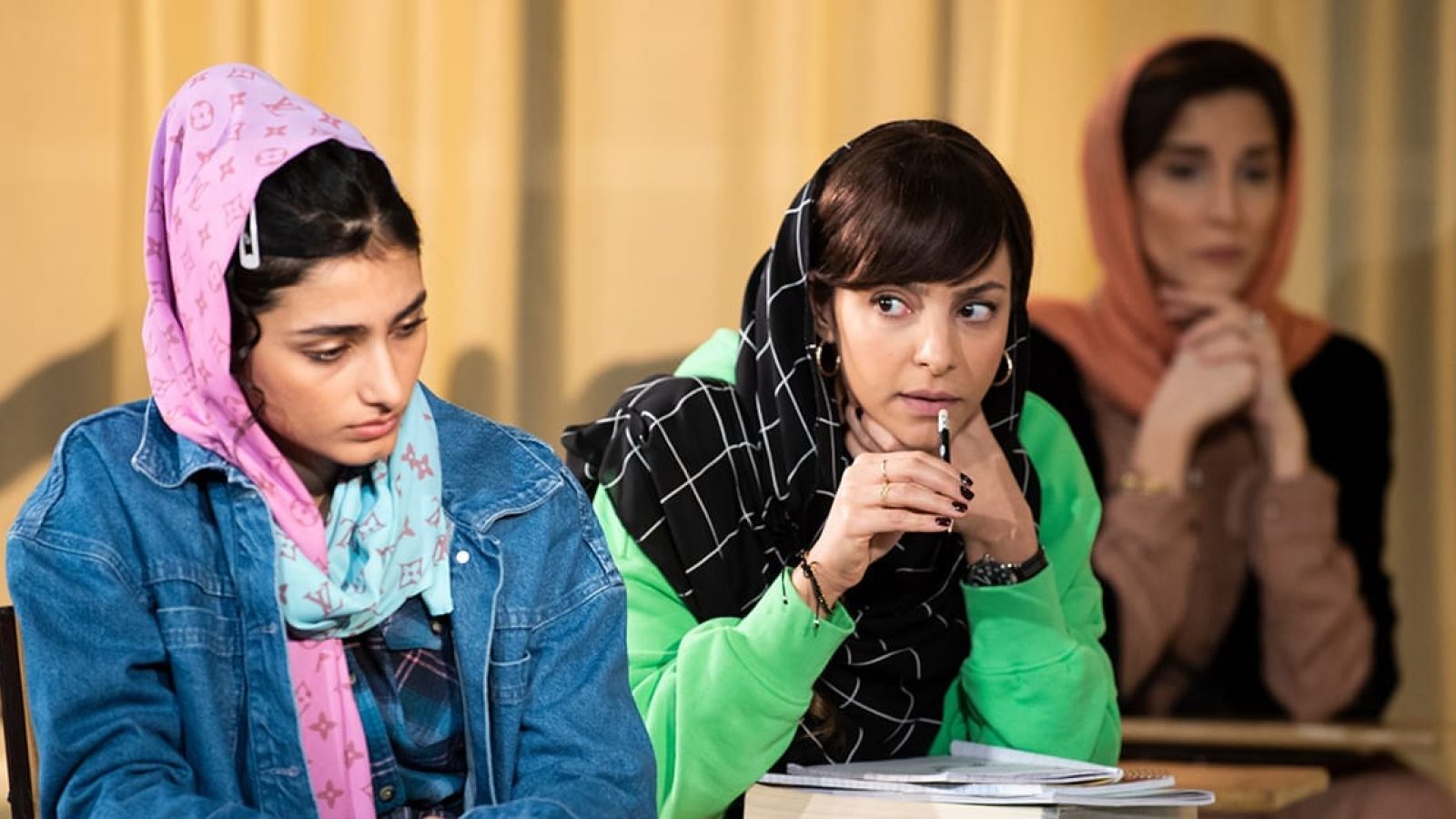 Three women in hijabs sit at desks, one thoughtfully holding a pen.