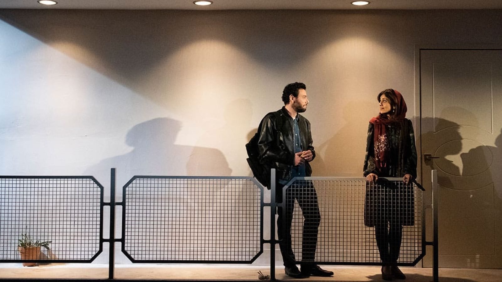 Man and woman converse on a balcony, separated by a mesh fence.