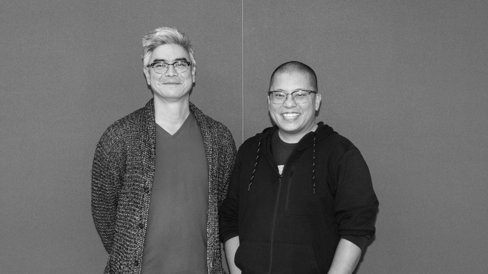 Two men stand shoulder to shoulder against a gray background. One wears a cardigan, the other a zip-up jacket.