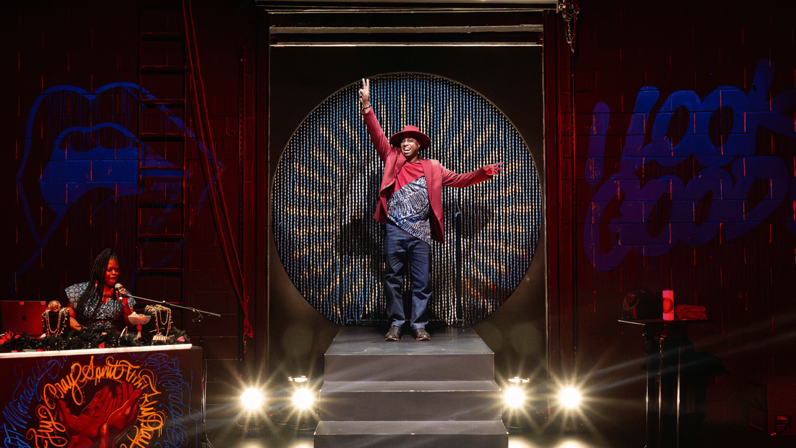 A performer in a red hat and jacket on stage, arms raised. DJ on the left.