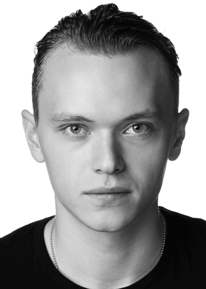 GILLES-GEARY Headshot of a young man with short, slicked-back hair, wearing a black t-shirt and a silver chain.
