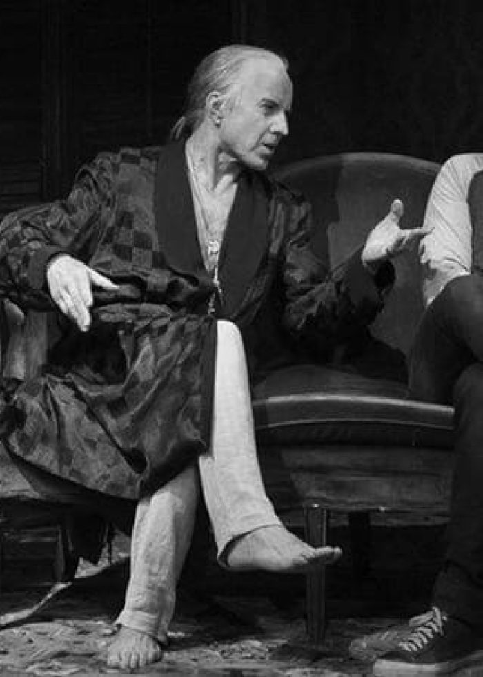 Actors in a scene from "Ghost Stories: The Shawl and Prairie du Chien" play. Man in robe talks to another on a couch.