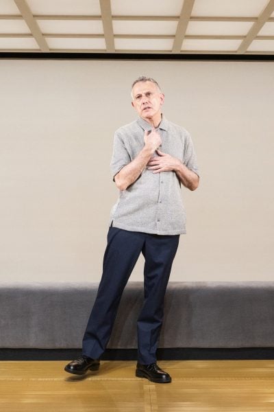 David Greenspan, standing, wearing a gray shirt and dark pants.