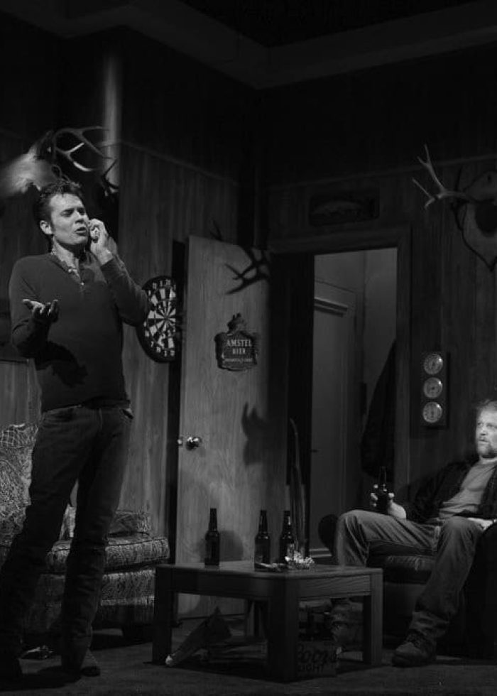 Two men in a dimly lit room from Kenneth Lonergan's 'Hold On to Me Darling,' featuring hunting trophies and beer.