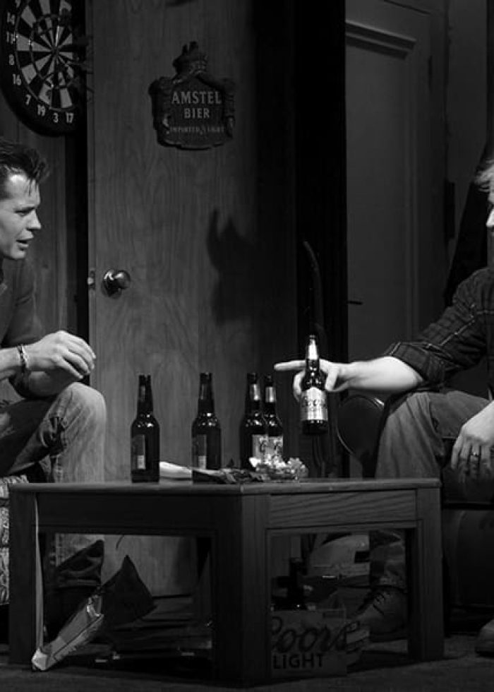 Two men in a scene, possibly from a play, with beer bottles. Dimly lit room.
