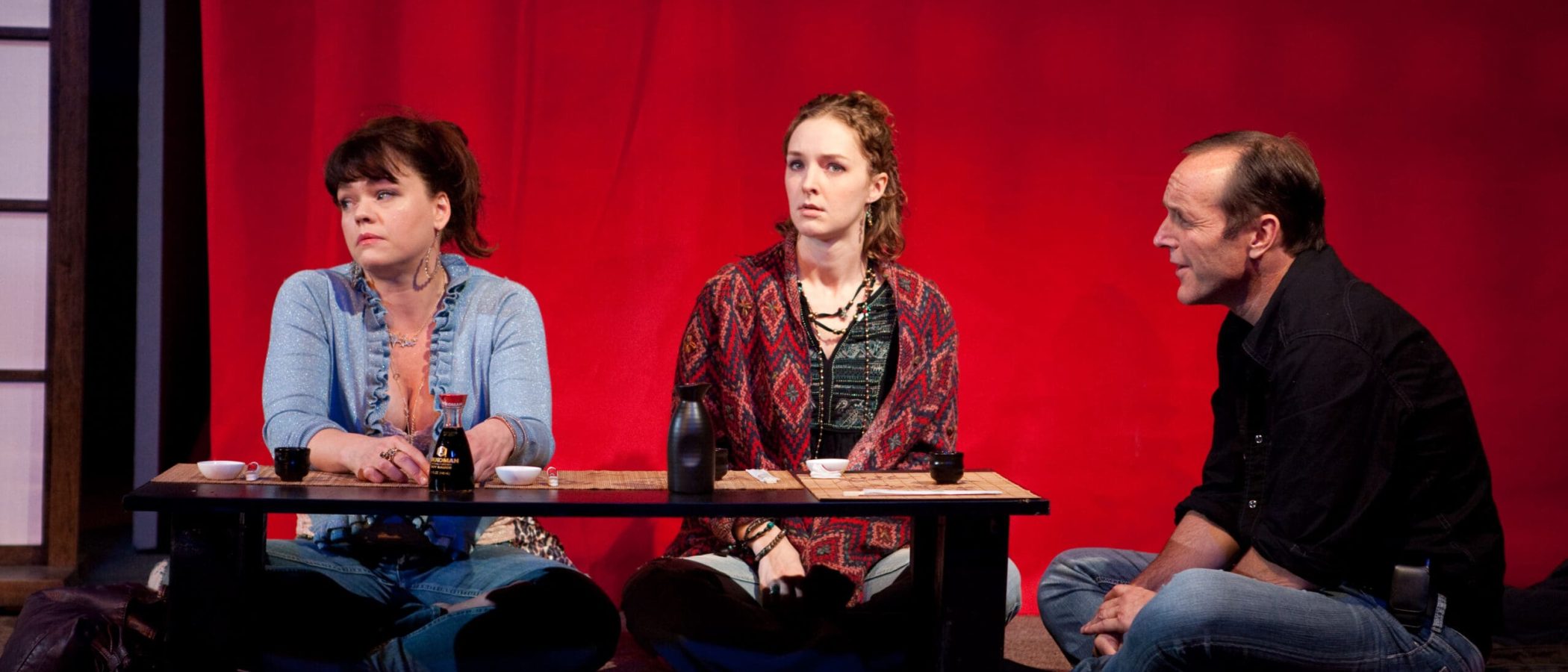 Actors in a play seated at a table on stage with a red backdrop. Soy sauce bottle visible.