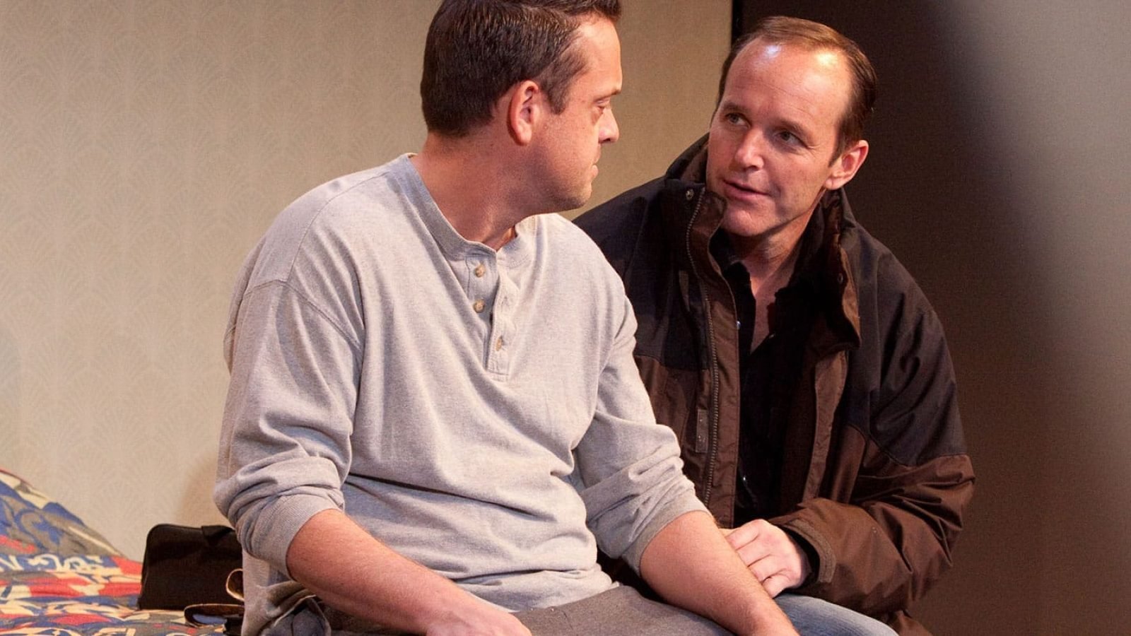 Two men share a quiet moment, seated near a bed.