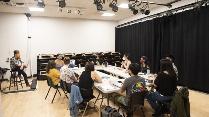 Group meeting in a rehearsal space with stage lights and people wearing masks.