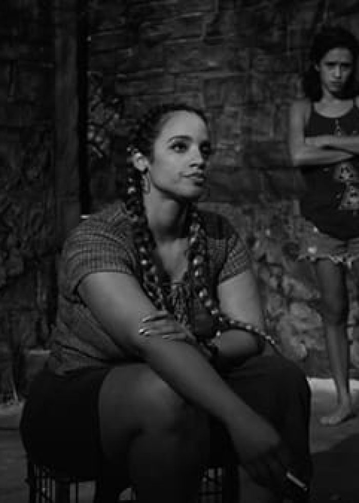 Two women in a scene from the play 'Tell Hector I Miss Him'. One seated, the other standing, both in a dimly lit room.