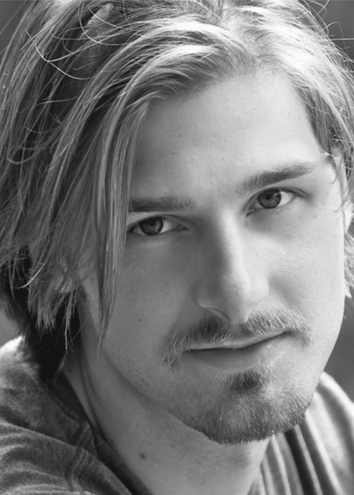 Close-up portrait of a young man with medium-length blonde hair and a goatee, looking at the camera.