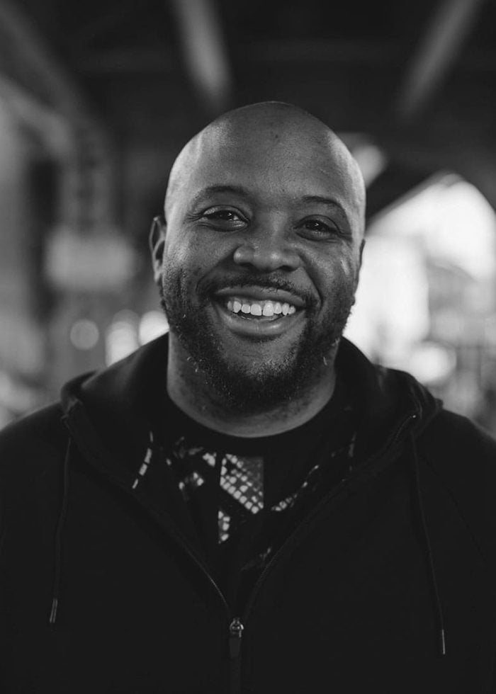 Idris Goodwin smiling, wearing a black hoodie, looking at the camera.