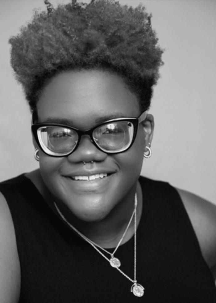 Ibi Owolabi smiling, wearing glasses and a necklace, looking at the camera