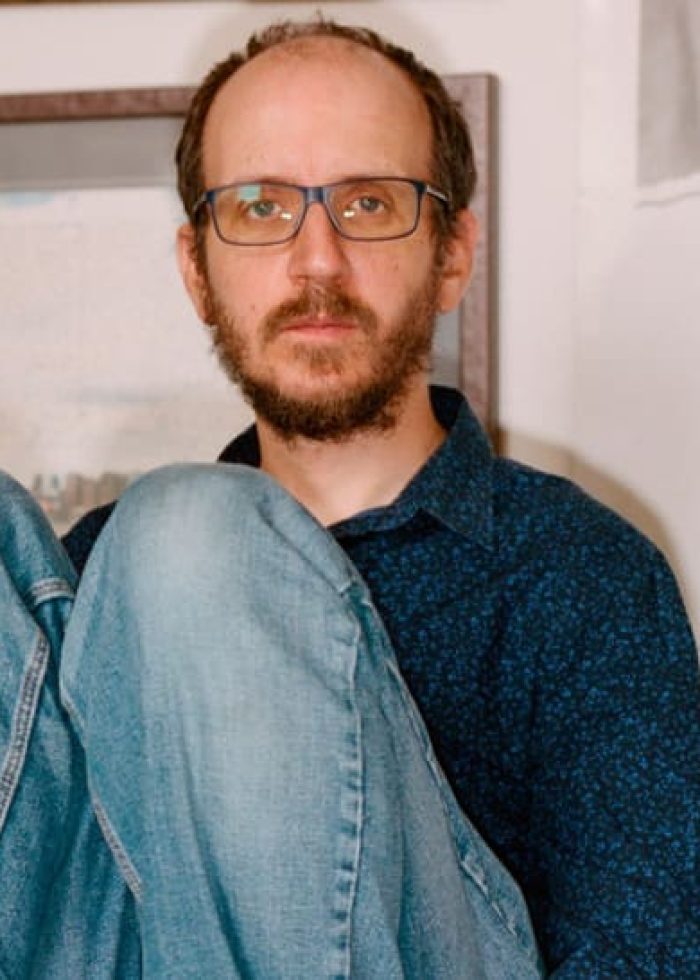 Man with glasses sitting on a couch, surrounded by framed art and a poster.