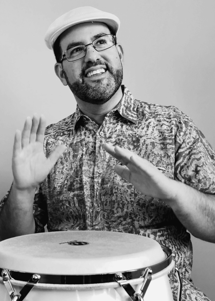 Man playing conga drum, smiling. Wearing a hat and glasses while drumming.