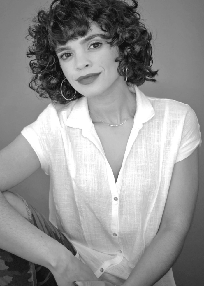 Gizel Jiménez, a woman with curly hair, wearing a white shirt and hoop earrings, smiles at the camera.