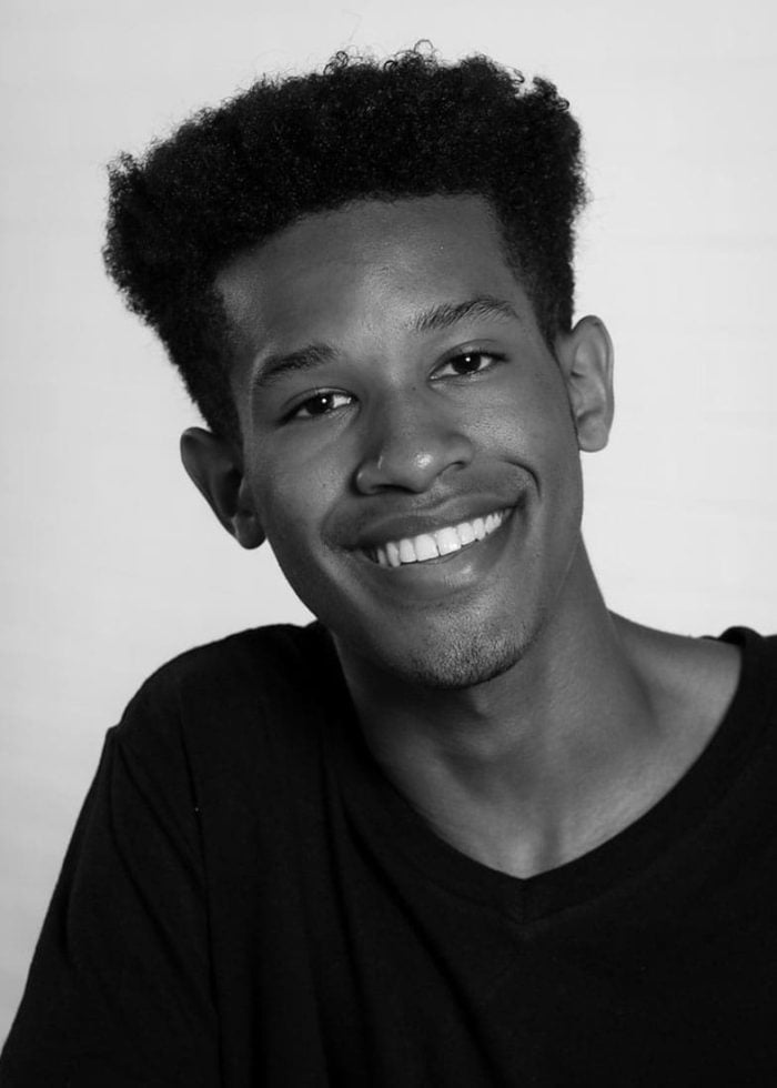 Smiling young man with afro, wearing a black V-neck shirt.