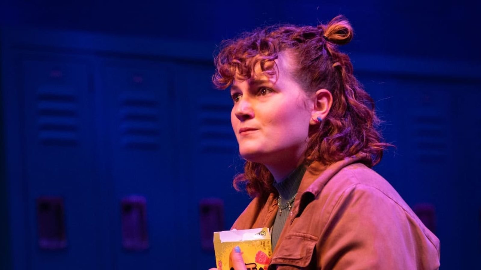 A young woman with curly hair holds a juice box, looking thoughtfully.