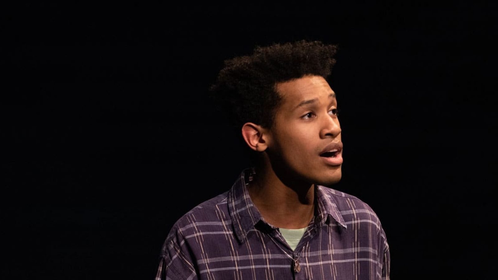 Young man in plaid shirt, looking upward, possibly in a theatrical performance.