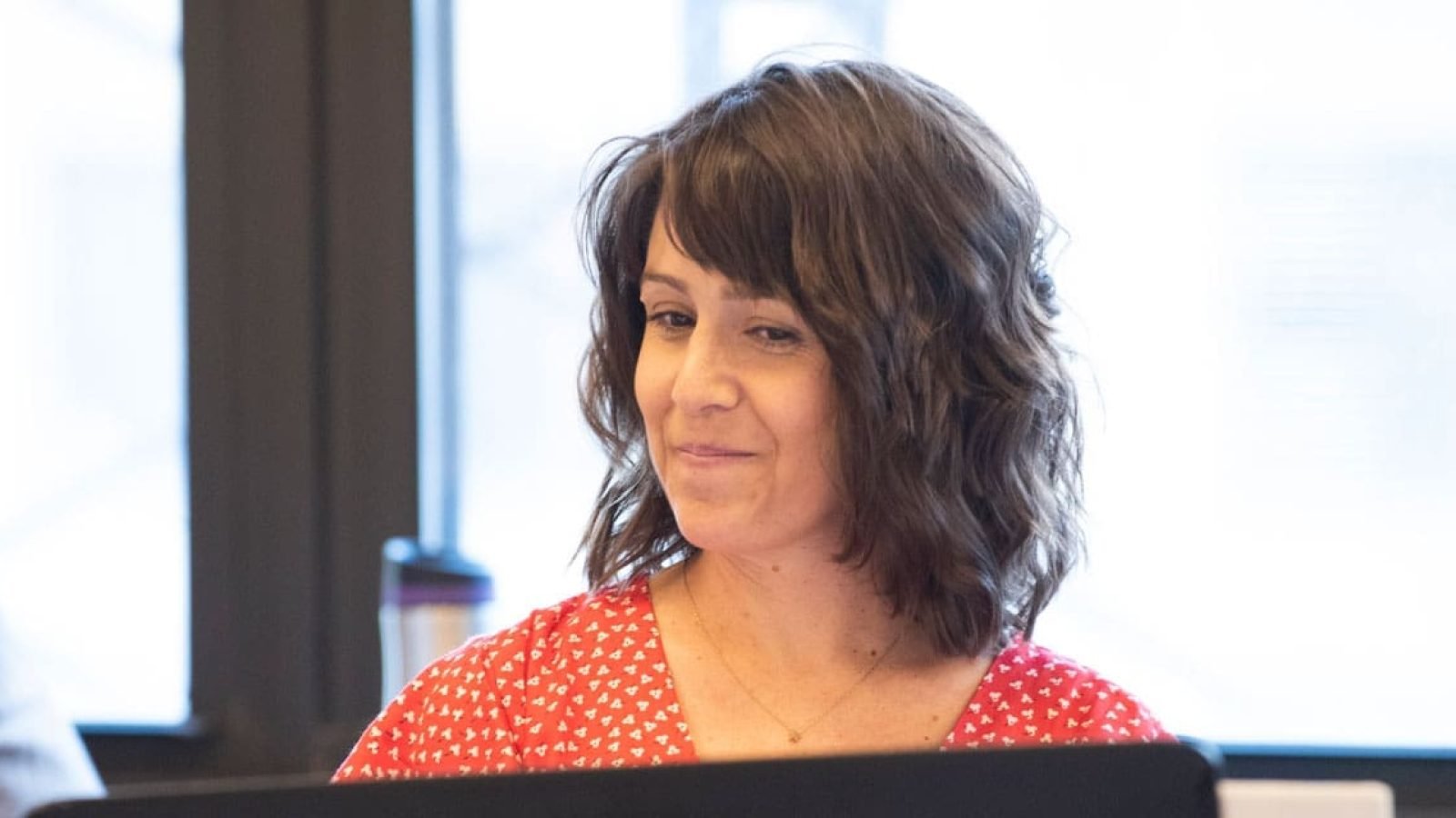Woman with short brown hair, wearing a red floral dress, sits at a table.