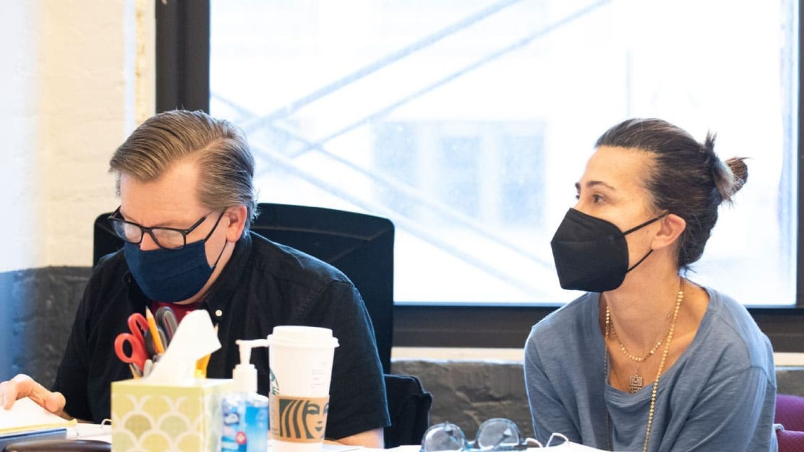 Two masked individuals, likely involved in a theatrical production, review paperwork at a table.