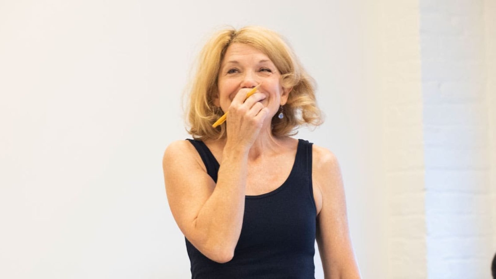 Smiling woman with blonde hair, holding a pencil to her mouth, wearing a black tank top.