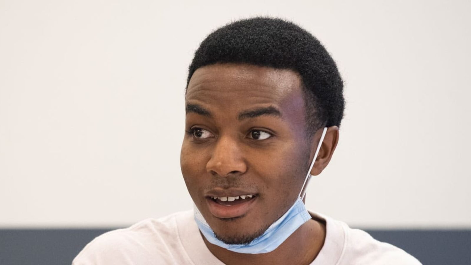 Close-up portrait of a young Black man wearing a light-colored shirt and a light blue face mask.