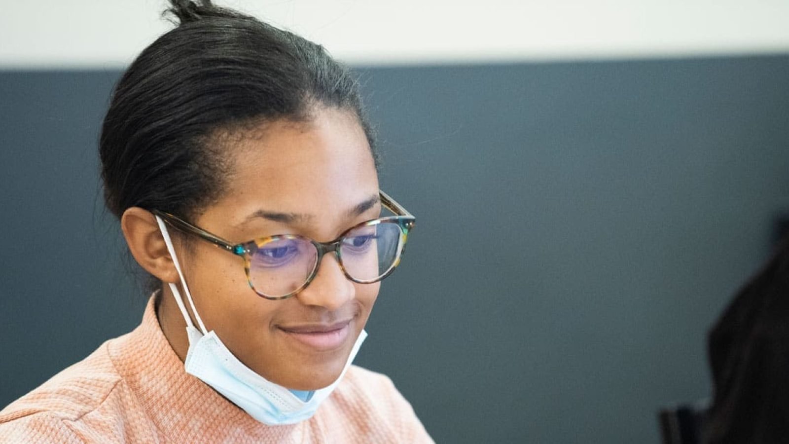 Young woman wearing glasses and a face mask, looking down.