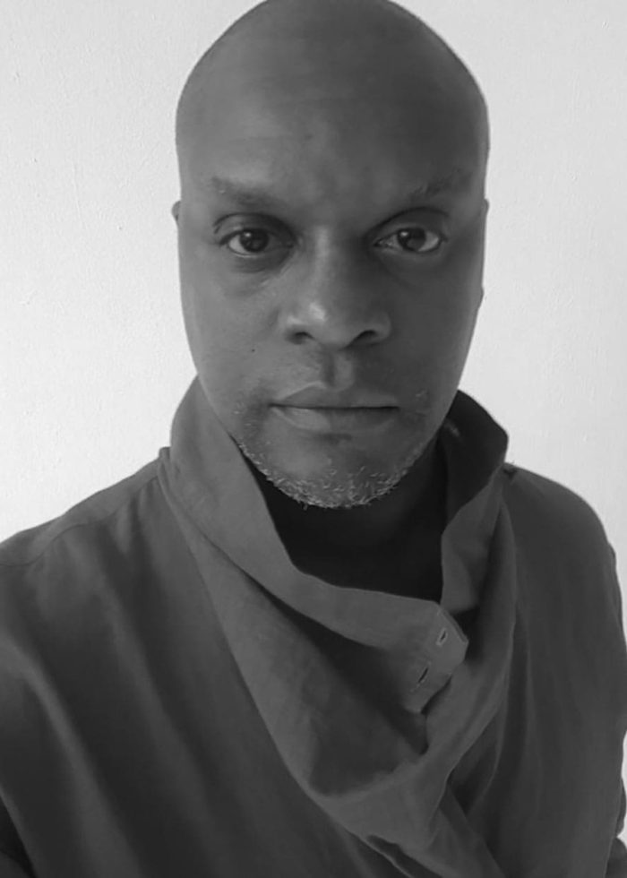 Close-up of Kevin Mambo, bald man with a serious expression, wearing a draped collar shirt.