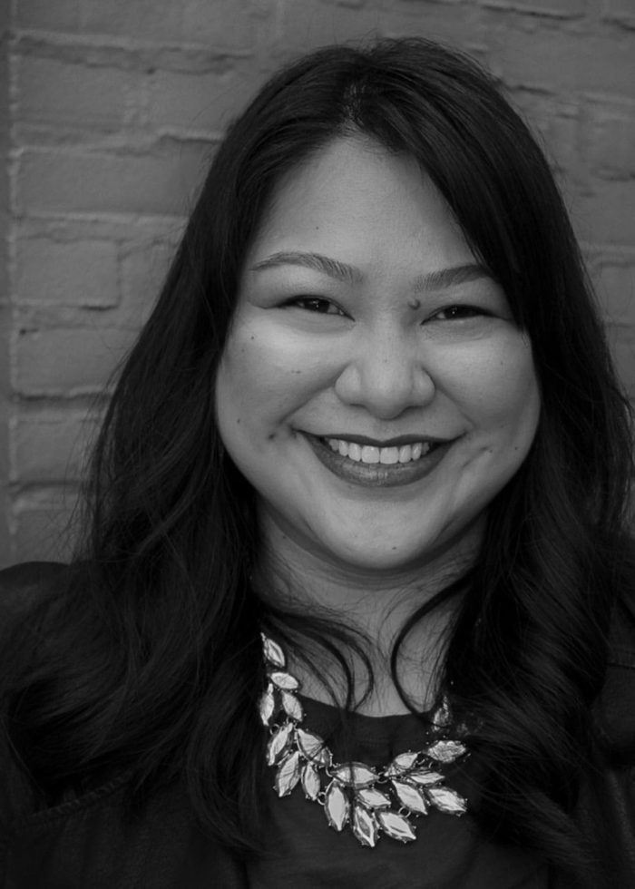 Kristi Donna Ng smiling at the camera, wearing a statement necklace, in front of a brick wall